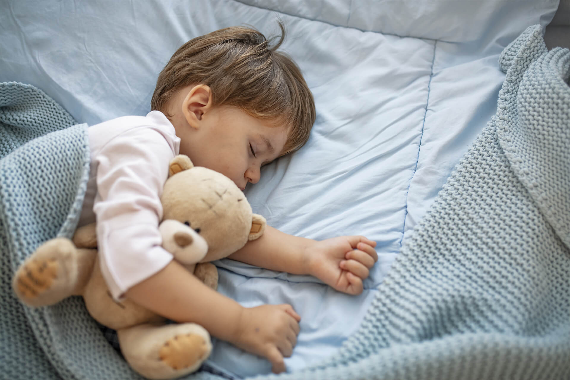 Kleines Kind liegt Schlafen in einem blau bezogenen Bett. Dabei kuschelt das Kind mit einem beigefarbenen Teddy im Arm.