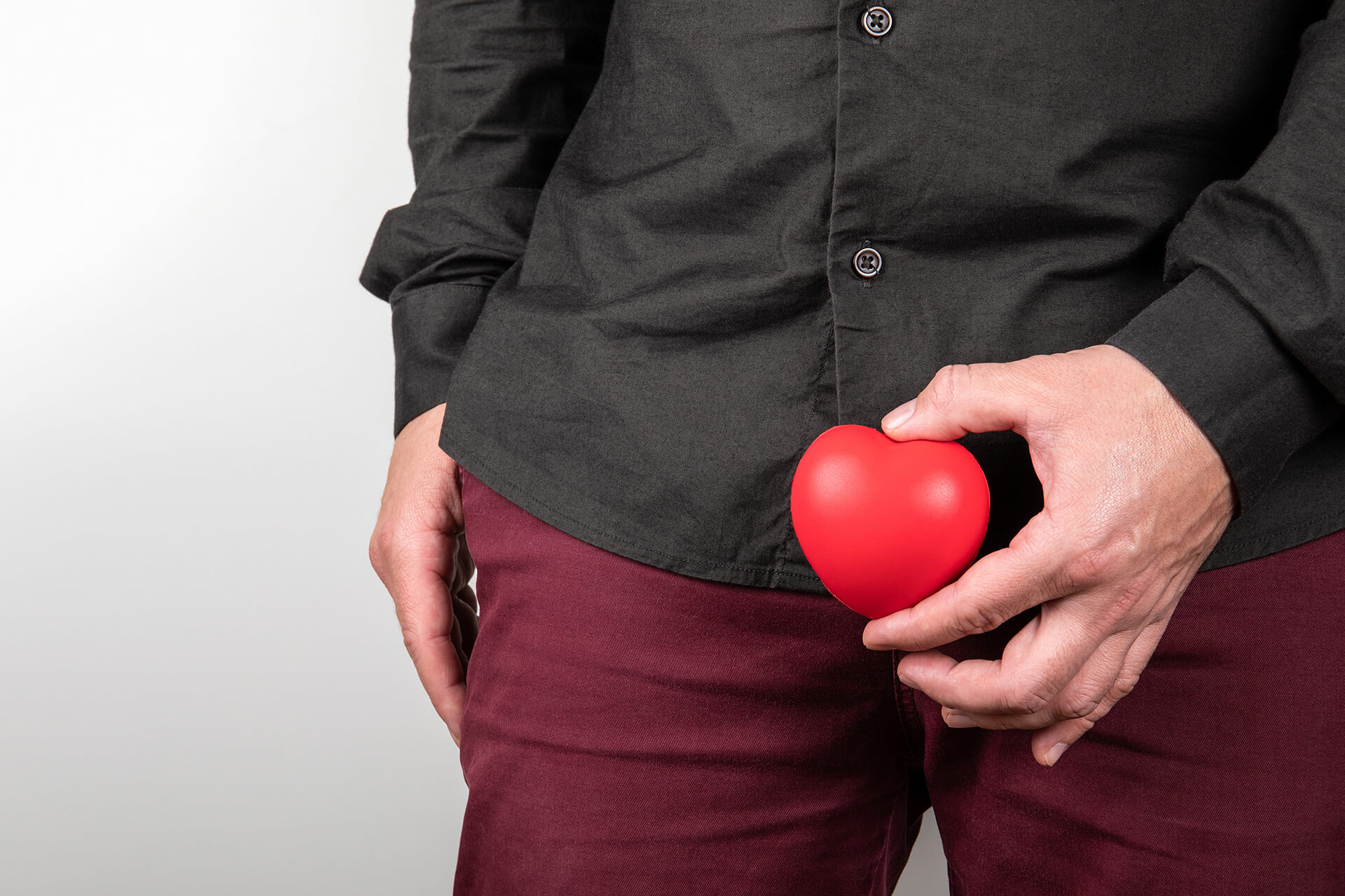Man sieht den Unterleib von einem Mann in roter Hose und schwarzen Hemd. Vor dem Intimbereich hält er ein kleines rotes Herz aus Kunststoff in Rot.