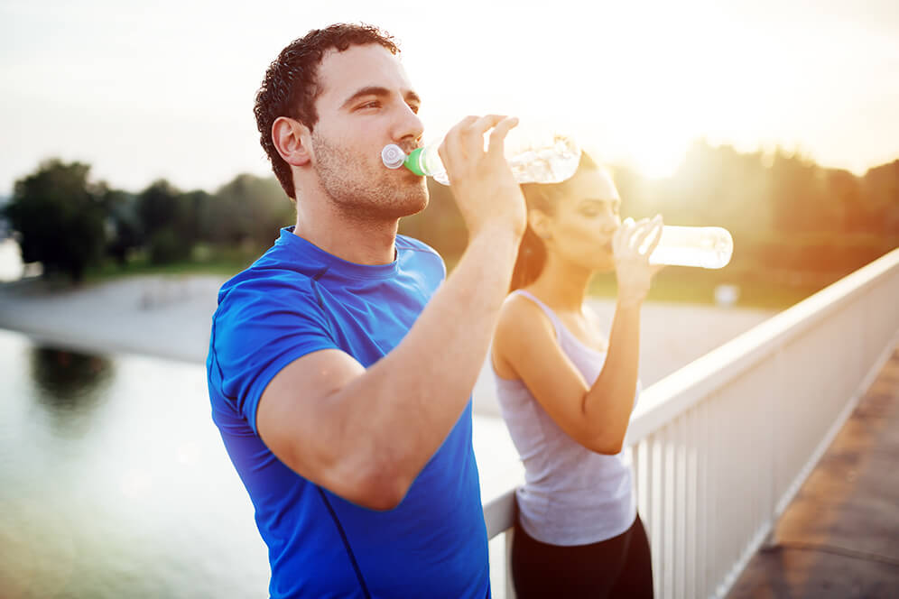 Ein Mann und eine Frau, beide sportlich gekleidet, trinken gemeinsam viel Wasser aus einer Flasche auf einer Brücke, während im Hintergrund die Sonne aufgeht.